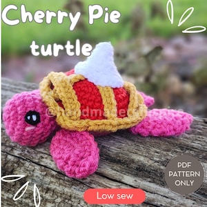 以下が含まれることがあります： ピンクの体と背中にチェリーパイの殻をつけた、かぎ針編みのかめのおもちゃ。パイの殻は赤と黄色で、白いホイップクリームのトッピングがされています。おもちゃは木製の表面の上に置かれています。画像には「Cherry Pie turtle」と「Low sew」の文字が見えます。