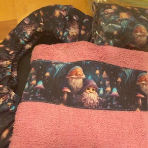 May include: A pink towel with a decorative band featuring a whimsical illustration of gnomes and mushrooms. A matching dark blue fabric item and a small pillow with the same gnome and mushroom design are also present.