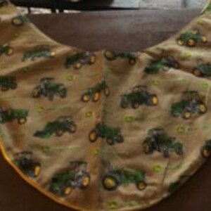May include: A green and yellow fabric baby bib with a pattern of John Deere tractors. The bib is shaped like a crescent moon and has a yellow trim.
