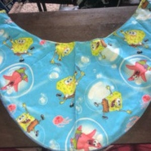 May include: A light blue, crescent-shaped fabric bag featuring a repeating pattern of cartoon characters, including a yellow sponge and a pink starfish, set against a background of bubbles. The bag is likely handmade and suitable for carrying small items.