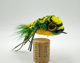 Può includere: Un'esca da pesca a mosca artigianale a forma di pesce, con dettagli gialli, verdi e neri. L'esca è appoggiata su un tappo di sughero con la scritta "APOTHIC WINEMAKER'S". L'esca ha un occhio dettagliato e lunghe fibre verdi.