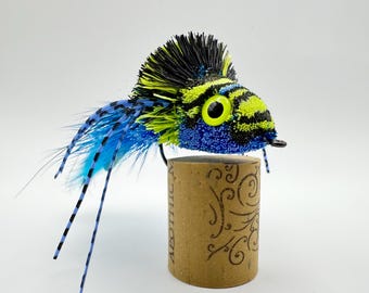 Può includere: Un'esca da pesca colorata, blu, nera e gialla, posizionata su un tappo di sughero decorativo. L'esca presenta una testa dettagliata e lunghe estensioni piumate. Perfetta per i pescatori, è progettata per attirare i pesci.