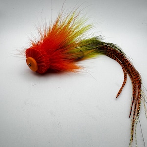 Puede incluir: Un señuelo de pesca con mosca que presenta una cabeza de espuma de color naranja brillante con un ojal metálico central, rodeada de fibras sintéticas naranjas y amarillas, y una cola larga compuesta por plumas barradas marrones y negras.