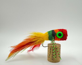 Può includere: Un'esca da pesca colorata con un design rosso, arancione, giallo e verde. L'esca ha un grande occhio verde e un amo. L'esca è fatta di piume e altri materiali. L'esca è appoggiata su un tappo di sughero.