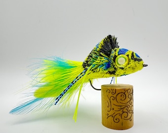 Può includere: Un'esca da pesca a mosca vibrante e fatta a mano. L'esca presenta un corpo giallo con dettagli neri e blu e lunghe piume fluenti nelle tonalità del giallo e del blu. Accanto all'esca c'è un piccolo tappo decorativo con un disegno inciso.