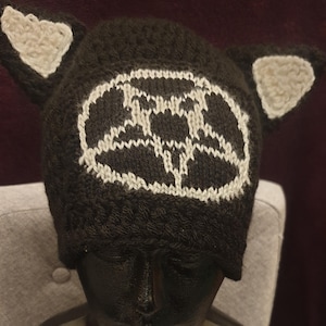 May include: A brown knitted hat with white knit ears and a white knit design on the front. The design is a circle with a pattern of lines and dots.