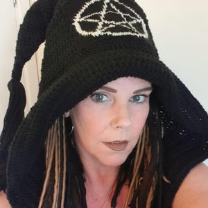 Puede incluir: Sombrero de ganchillo negro con capucha puntiaguda y diseño de pentagrama blanco. El sombrero está hecho de un hilo grueso y texturizado y tiene una borla larga y colgante. El sombrero lo lleva una persona con rastas.