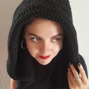 May include: A black crocheted hood with a textured pattern. The hood is worn over the head and shoulders. The person is wearing a black top and has silver hoop earrings.