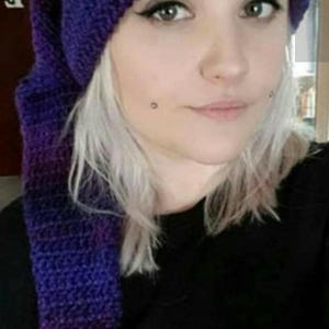 May include: A long, slouchy crochet beanie in a deep purple and blue variegated yarn. The hat features a textured, hand-stitched pattern with a long, trailing tail that hangs down the side, crafted from soft, thick yarn in dark, blended tones.