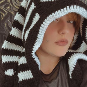 Puede incluir: Una bufanda con capucha de crochet a rayas blancas y negras con un diseño de círculo negro de crochet en la capucha.