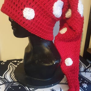 Puede incluir: Un sombrero rojo de ganchillo hecho a mano con lunares blancos y una larga cola puntiaguda. El sombrero se exhibe en una cabeza de maniquí negra. La cola termina en una borla. El sombrero es un accesorio caprichoso.