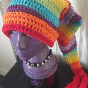 Puede incluir: Un gorro de ganchillo hecho a mano con colores del arcoíris, incluyendo rojo, naranja, amarillo, verde, azul y morado. El gorro tiene una larga cola puntiaguda con una borla roja. Se muestra en una cabeza de maniquí morada.