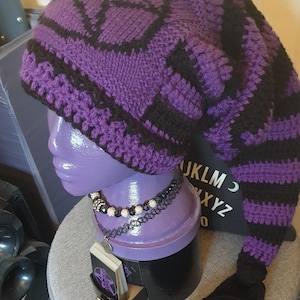 May include: Purple and black knitted hat with a long black tassel. The hat has a geometric pattern and a long tail. The hat is displayed on a purple mannequin head.