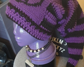 May include: Purple and black knitted hat with a long black tassel. The hat has a geometric pattern and a long tail. The hat is displayed on a purple mannequin head.
