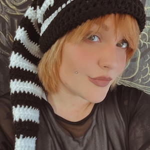 May include: A black and white striped crochet hat with a long tail. The hat has a black brim and a white stripe around the top. The tail is long and has a black tassel at the end.