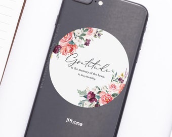 May include: A black iPhone with a white sticker featuring a floral design and the quote "Gratitude is the memory of the heart." by Mary Mackillop.