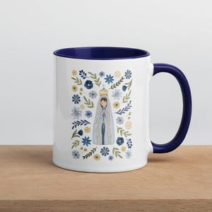 Op de afbeelding: Witte keramische mok met een blauwe rand en handvat. De mok heeft een bloemenmotief met een afbeelding van de Maagd Maria in het midden. De bloemen zijn blauw, geel en groen.