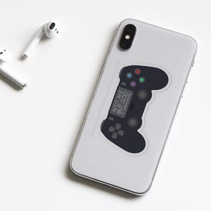 May include: A white iPhone with a black PlayStation controller sticker. The sticker has the text "NOT ME BUT GOD" and "ST. JAMES 2:26" on it.