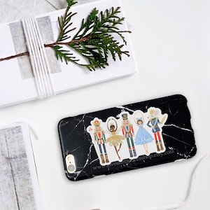 May include: A black phone case with a white marble design features a sticker of a Nutcracker and a ballerina. The sticker is colorful and includes a mouse and a crown. The text "www.sweetlittlethingshop.etsy.com" is printed on the case.