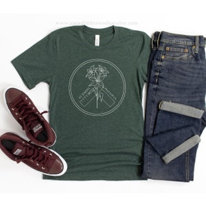 May include: A dark green t-shirt with a white circular graphic featuring a flower and the text "ST. JOSEPH PRAY FOR US." The shirt is paired with blue jeans and maroon sneakers.