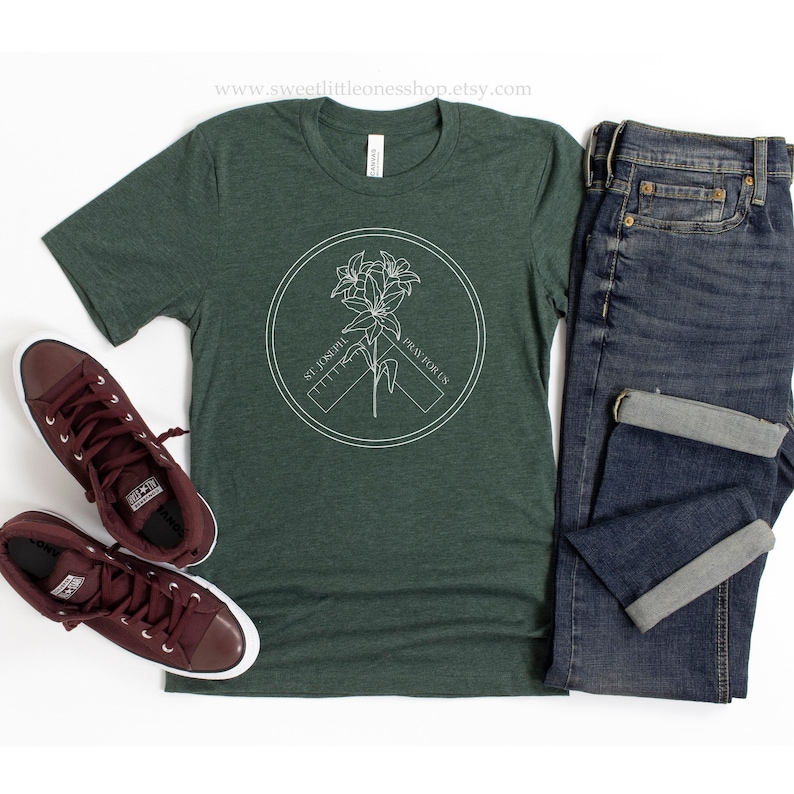May include: A dark green t-shirt with a white circular graphic featuring a flower and the text "ST. JOSEPH PRAY FOR US." The shirt is paired with blue jeans and maroon sneakers.