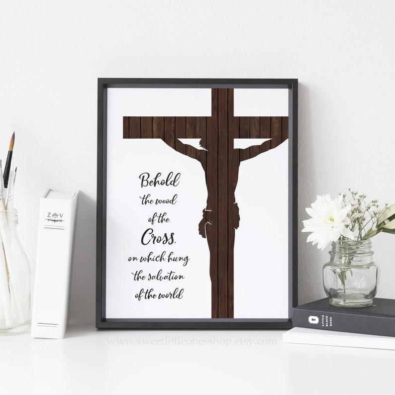May include: A black framed print with a white background and a brown wooden cross silhouette. The cross has a silhouette of a person on it. The text on the print reads "Behold the wood of the Cross. On which hung the salvation of the world." The website address "www.sweetlittleonesshop.etsy.com" is printed at the bottom of the print.