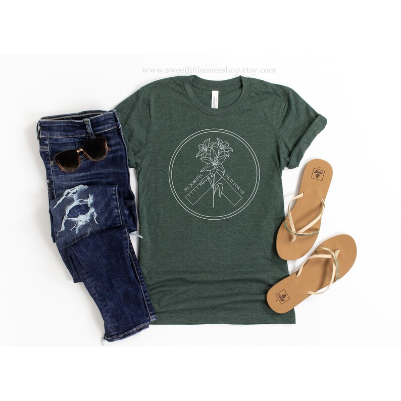 May include: Dark green t-shirt with a white circular graphic featuring a floral design and the text "ST. JOSEPH, PRAY FOR US." The shirt is paired with blue jeans, sunglasses, and tan sandals.