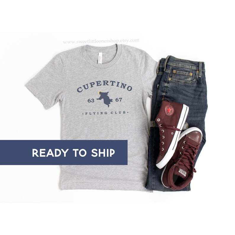May include: A heather gray t-shirt with the text "Cupertino Flying Club" and a map graphic, paired with dark wash jeans and maroon high-top sneakers. The words "Ready to Ship" are displayed on a blue banner.