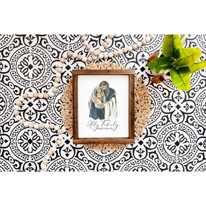 May include: A framed watercolor painting of the Holy Family with the text "Holy Family Pray For Us". The painting is on a brown wooden frame and is sitting on a woven straw mat.