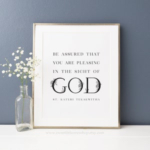 Op de afbeelding: Een zwart-witte printbare wandkunstprint met een bloemmotief rond het woord "GOD" en de tekst "WEES ERVAN OVERTUIGD DAT JE BEHAGELIJK BENT IN DE OGEN VAN GOD ST. KATERI TEKAKWITHA".