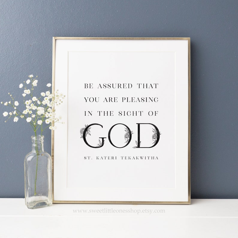 Op de afbeelding: Een zwart-witte printbare wandkunstprint met een bloemmotief rond het woord "GOD" en de tekst "WEES ERVAN OVERTUIGD DAT JE BEHAGELIJK BENT IN DE OGEN VAN GOD ST. KATERI TEKAKWITHA".