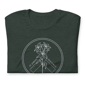 May include: Dark green t-shirt with a white circular graphic. The graphic features a lily flower design with the text "ST. JOSEPH. PRAY FOR US" in a circular pattern. The t-shirt is folded.