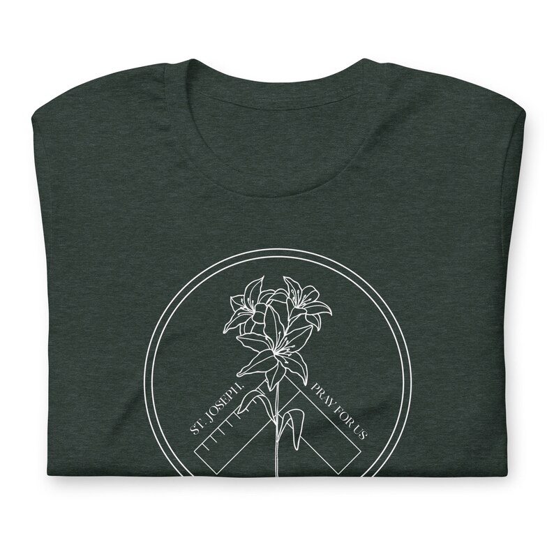 May include: Dark green t-shirt with a white circular graphic. The graphic features a lily flower design with the text "ST. JOSEPH. PRAY FOR US" in a circular pattern. The t-shirt is folded.