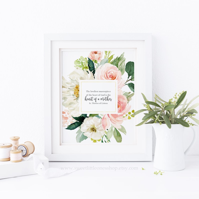 May include: A white framed print featuring a watercolour floral design in shades of pink and white. The print includes a quote by St. Th&eacute;r&egrave;se of Lisieux: "The loveliest masterpiece of the heart of God is the heart of a mother."
