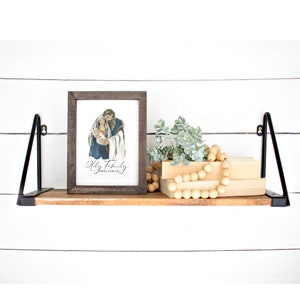 May include: A framed print of a watercolor painting of the Holy Family. The painting depicts Mary, Joseph, and baby Jesus. The frame is brown wood and the print has the text "Holy Family Pray For Us".