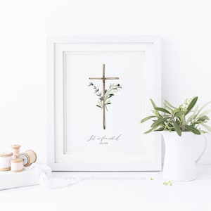 May include: A framed print featuring a watercolor illustration of a wooden cross adorned with olive branches and small purple flowers. The text "It is finished" and John 19:30 are written below the cross. The print is in a white frame.