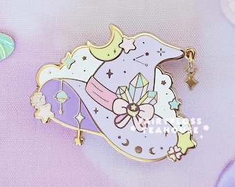 May include: A whimsical enamel pin in the shape of a witch's hat, adorned with celestial motifs, crystals, and stars. The pin is primarily lavender and gold, with a pink ribbon. The image also features a butterfly, flowers, and a bow, all in pastel colors.
