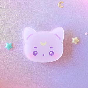 Pode incluir: Um pingente de acrílico em forma de gato lilás claro com um detalhe de lua crescente. O pingente tem olhos grandes e redondos e é cercado por pequenas estrelas e pérolas. O fundo é um gradiente de cores pastel com flores cor-de-rosa.