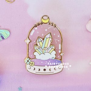 May include: A whimsical enamel pin featuring a gold-colored frame with a crystal cluster inside a glass dome. The pin is surrounded by decorative elements like stars, moons, and flowers, all set against a soft pink background.