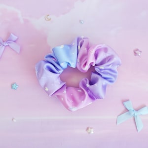 May include: A purple and blue ombre scrunchie with white stars and moons printed on it. The scrunchie is made of satin and is tied in a knot.