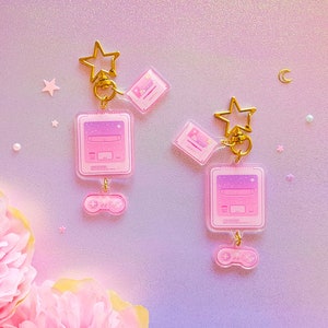May include: Two pink and purple video game console keychain charms with gold star charms and a pink and purple joystick charm. The charms are on a pink and purple background.