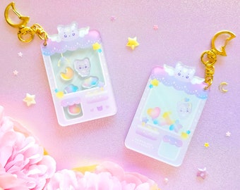 Peut inclure: Deux porte-clés en acrylique de couleur pastel avec un design fantaisiste. Chaque porte-clés présente un nuage avec un visage de chat et une petite fenêtre transparente avec un chat et des étoiles à l'intérieur. Le texte "A Magical Day" est imprimé au bas d'un porte-clés.