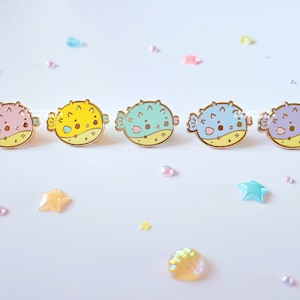May include: Five colorful enamel pins in the shape of pufferfish, each with a gold outline. The pins are in shades of pink, yellow, teal, blue, and purple. The background is white with scattered star and pearl-shaped decorations.
