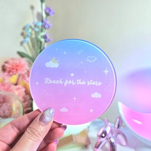 May include: A round, pink and blue gradient coaster with a white outline of clouds, stars, and a crescent moon. The text "Reach for the stars" is printed in white on the coaster.