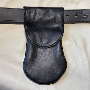 May include: Black leather pouch with a rounded bottom and a flap secured by a silver snap. The pouch is attached to a black leather belt. The background is a cream-colored surface.