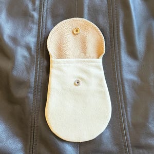 May include: A cream-colored leather pouch with a rounded bottom and a flap secured by a snap. The interior of the flap has a textured pattern. The pouch is open, revealing the interior. The background is a dark brown leather surface.