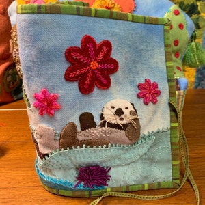 May include: A blue fabric pouch with a green and brown trim. The pouch features an embroidered sea otter, flowers, and a fuzzy purple pom-pom.