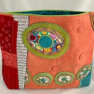 May include: A coral-colored zippered pouch with a patchwork design featuring a variety of fabrics and textures. The pouch has a green zipper and is decorated with colorful embroidered circles and flowers.