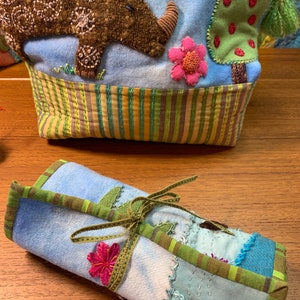 May include: A blue and green fabric roll with a green ribbon tied around it. The fabric has a patchwork design with different colors and textures. There is a pink flower embroidered on the fabric.