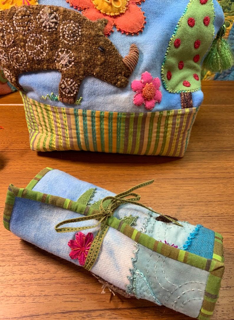 May include: A blue and green fabric roll with a green ribbon tied around it. The fabric has a patchwork design with different colors and textures. There is a pink flower embroidered on the fabric.